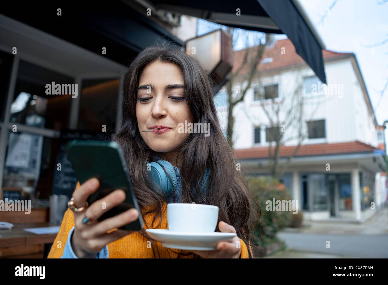 Femme tenant une cuillère dans la bouche et utilisant un téléphone intelligent près du café Banque D'Images Femme tenant une cuillère dans la bouche et utilisant un téléphone intelligent près du café Banque D'Images