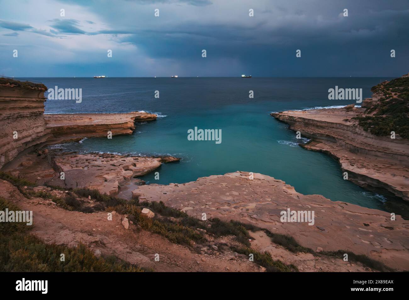 Vue sereine sur les falaises côtières de Malte à la piscine Peter's Pool, sous un ciel nuageux spectaculaire au crépuscule, avec des lumières de navire visibles au loin Banque D'Images