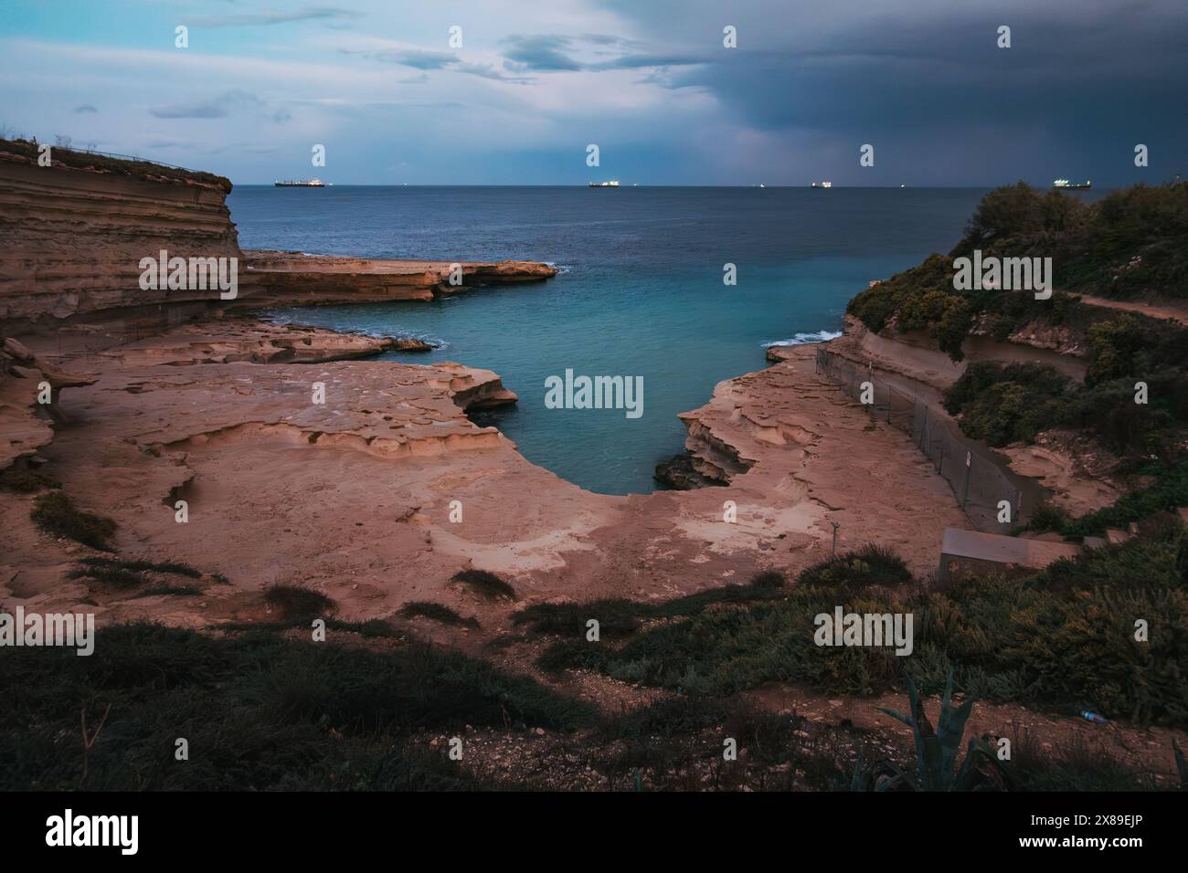 Vue sereine sur les falaises côtières de Malte à la piscine Peter's Pool, sous un ciel nuageux spectaculaire au crépuscule, avec des lumières de navire visibles au loin Banque D'Images