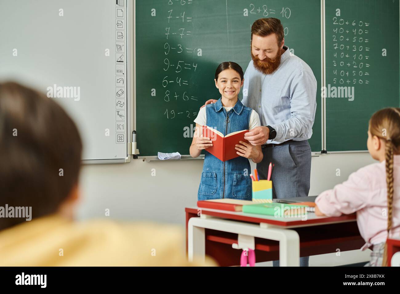 Un homme debout à côté d'une petite fille devant un tableau noir, enseignant dans une salle de classe lumineuse et animée. Banque D'Images