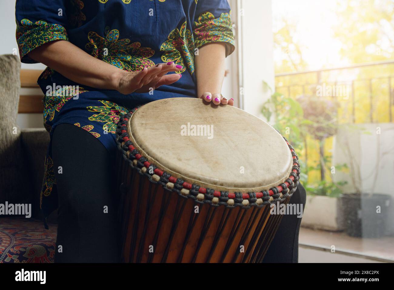 Mains féminines d'une femme méconnaissable jouant du tambour Djembe à la maison. Le tambour est marron et a un motif rouge et noir. La femme est dans un a détendu Banque D'Images