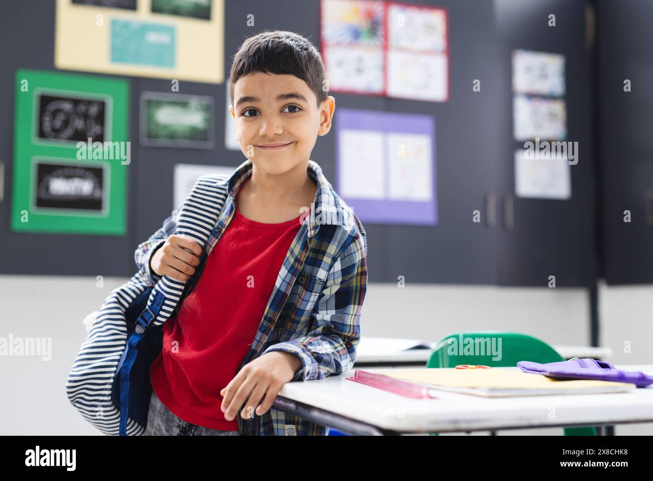 À l'école, un jeune garçon biracial avec un sac à dos se tient souriant dans la salle de classe, copiez l'espace Banque D'Images