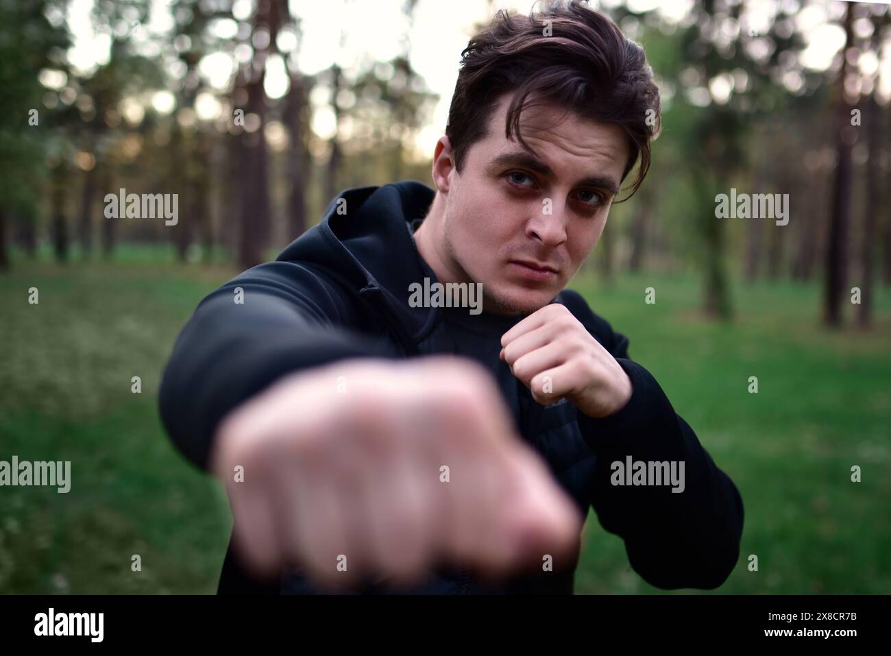 jeune homme sportif dans une veste noire sportive dans une pose de combat levant le poing pour un coup de poing sur un fond d'arbres forestiers Banque D'Images
