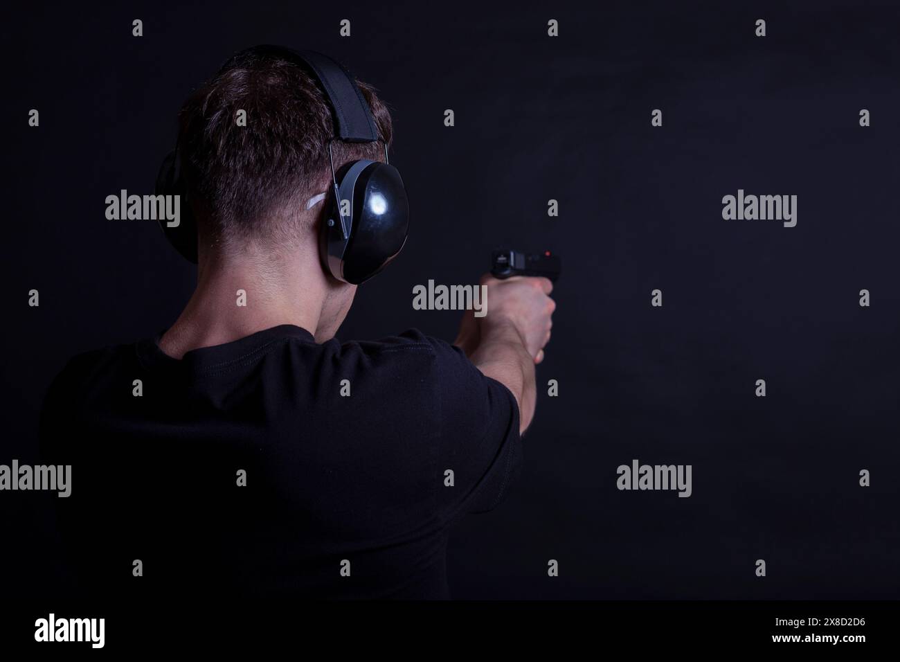 Jeune homme avec protection des oreilles et des yeux tirant un pistolet sur un fond noir Banque D'Images