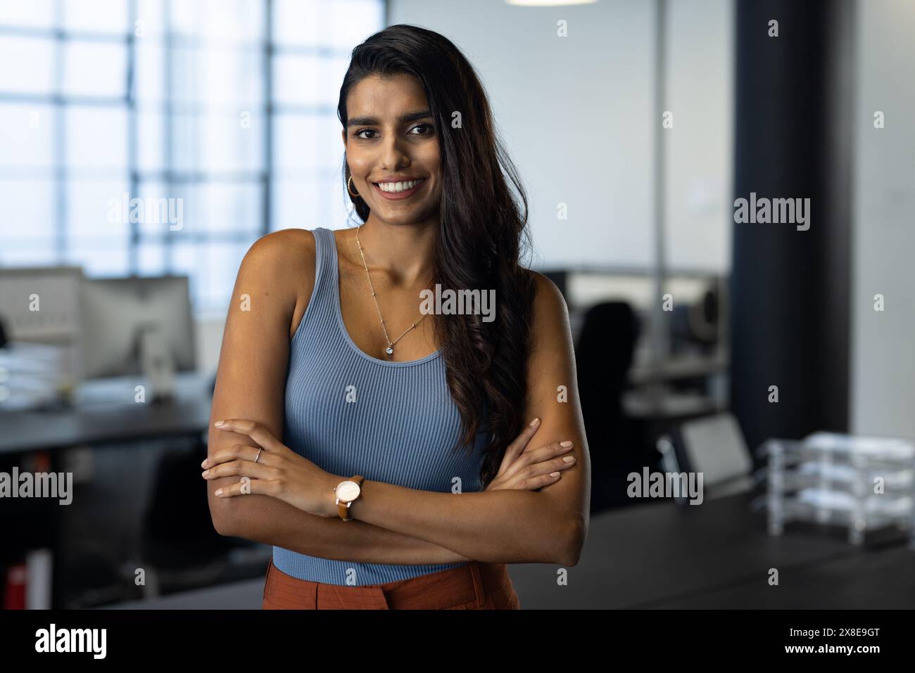 Au bureau moderne, la femme indienne se tient souriante, les bras croisés, en haut bleu sans manches. Elle a de longs cheveux foncés, des yeux bruns et un pantalon orange, avec calme Banque D'Images
