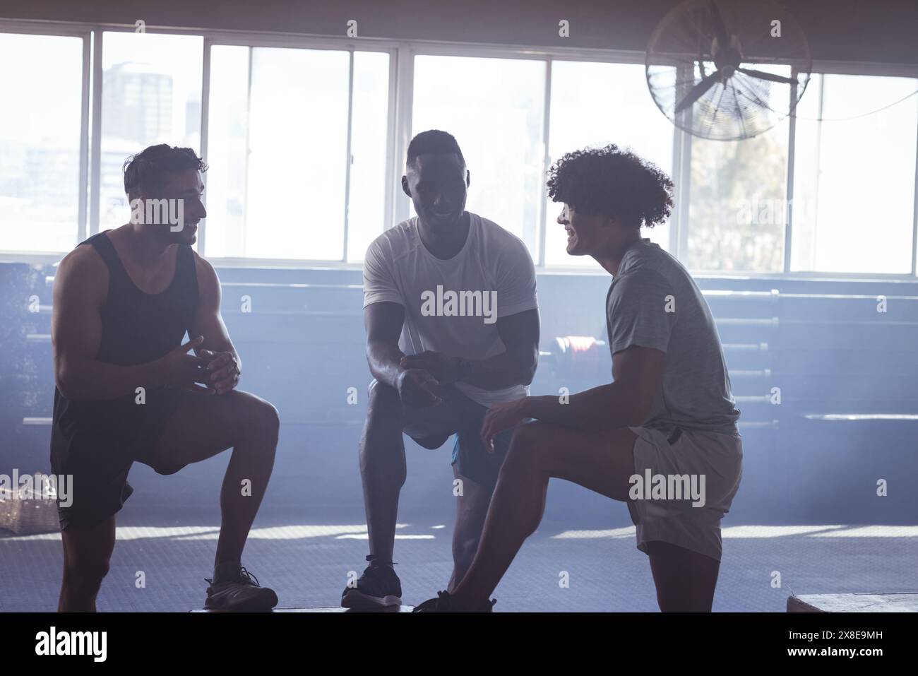 Trois jeunes hommes de toutes sortes en salle de gym discutant, prenant une pause de leur entraînement. La lumière du soleil traverse de grandes fenêtres, illuminant la salle de sport spacieuse Banque D'Images