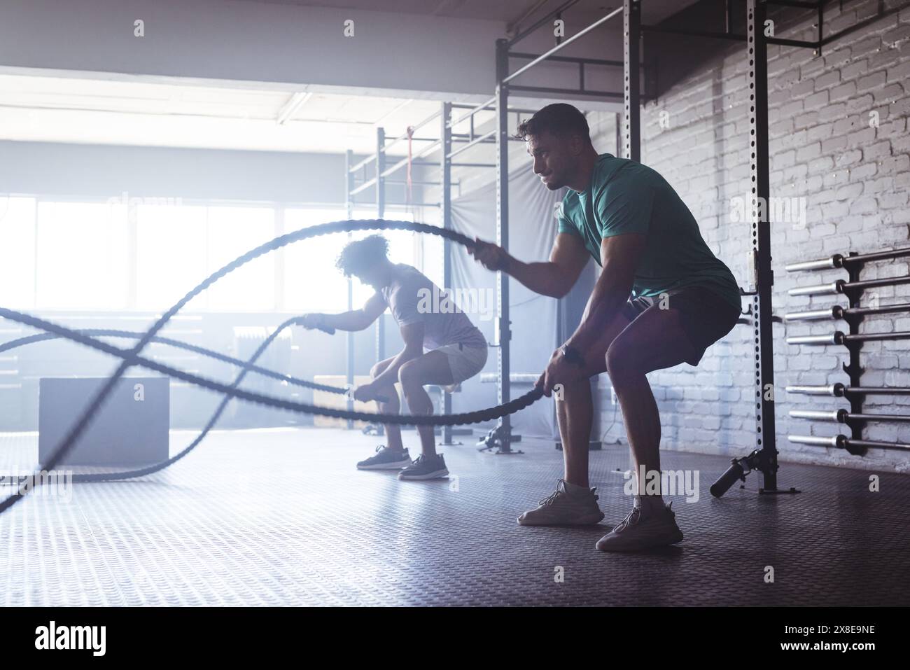 Deux hommes biraciaux utilisant des cordes de combat au gymnase, entourés par des hommes divers qui travaillent, copiez l'espace. Grandes fenêtres et divers appareils de fitness créant en Banque D'Images