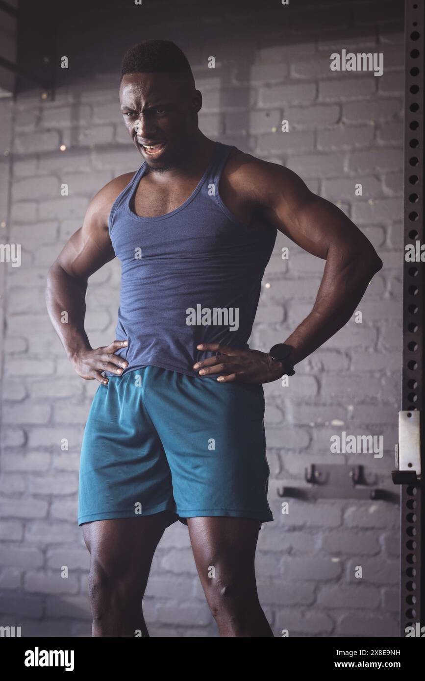 Homme afro-américain fort et en forme au gymnase portant un débardeur bleu et un short. Il est debout avec les mains sur les hanches contre fond de briques, regardant dissuasif Banque D'Images