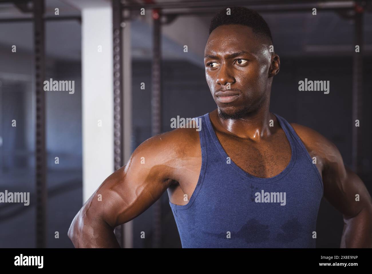 Homme afro-américain fort et en forme au gymnase dans le débardeur bleu, regardant déterminé. Debout dans une salle de gym moderne avec divers équipements d'entraînement en arrière-plan, Banque D'Images