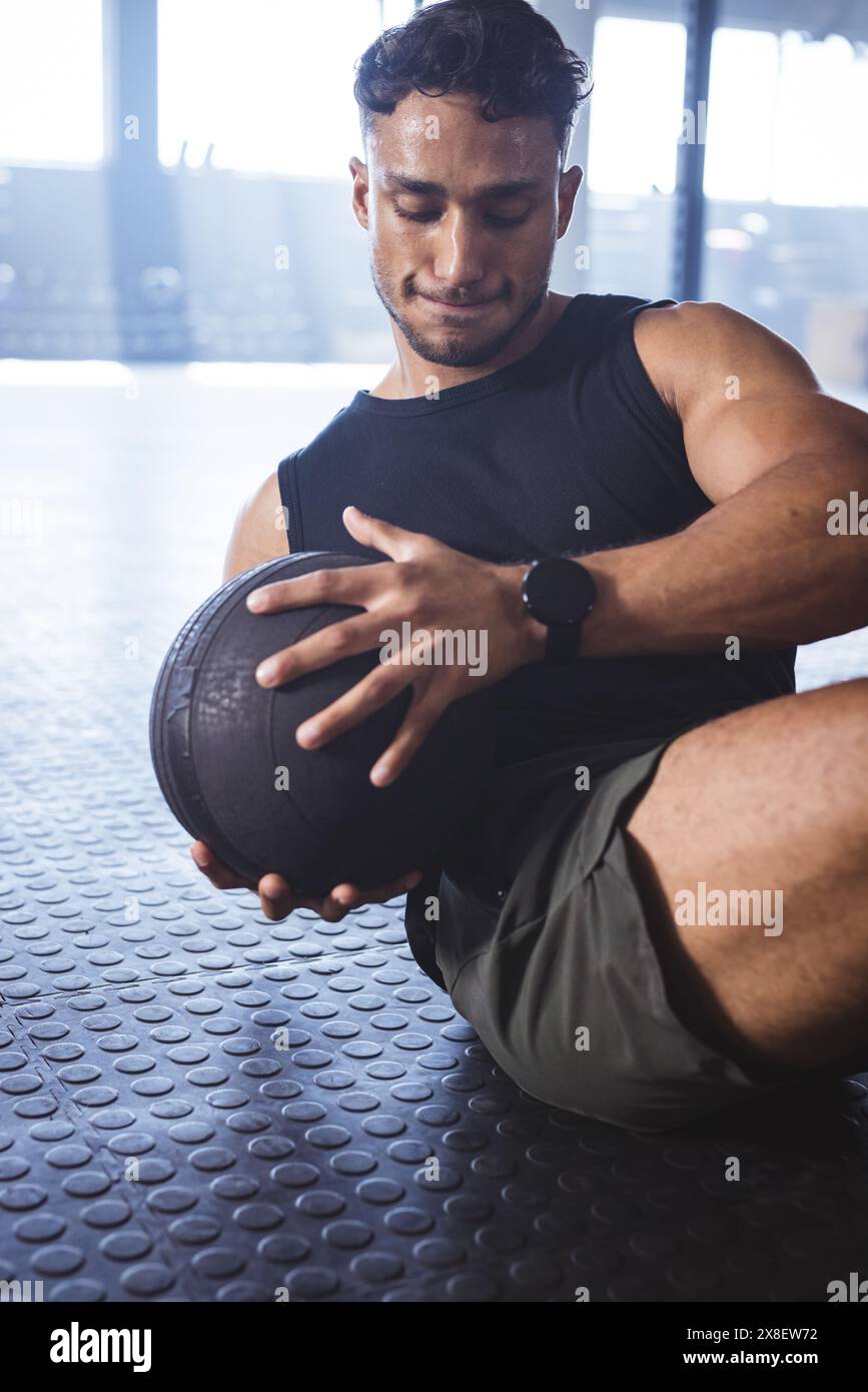 Homme biracial fort et en forme au gymnase tenant le medecine ball, s'entraînant. Sol de gymnastique en caoutchouc avec équipement de gymnastique en arrière-plan, poids et machines proprement Banque D'Images