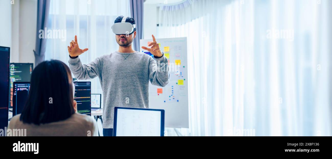 Un développeur de logiciels asiatique portant un casque de réalité virtuelle travaille sur un projet VR, avec une collègue concentrée sur son ordinateur Banque D'Images