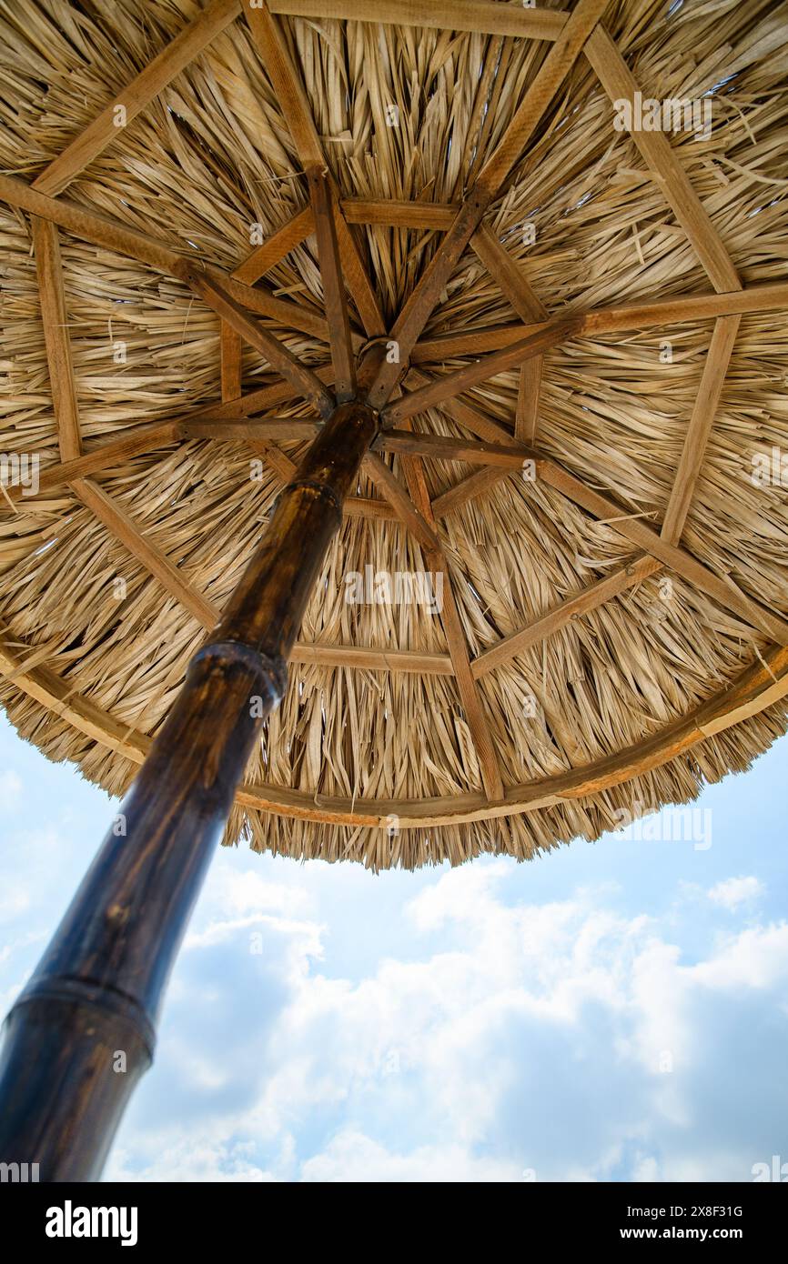 Un regard vers le haut révèle la symétrie complexe et la texture naturelle d'un parasol de plage en paille, encadré par le bleu serein d'un ciel moucheté de nuages. Banque D'Images