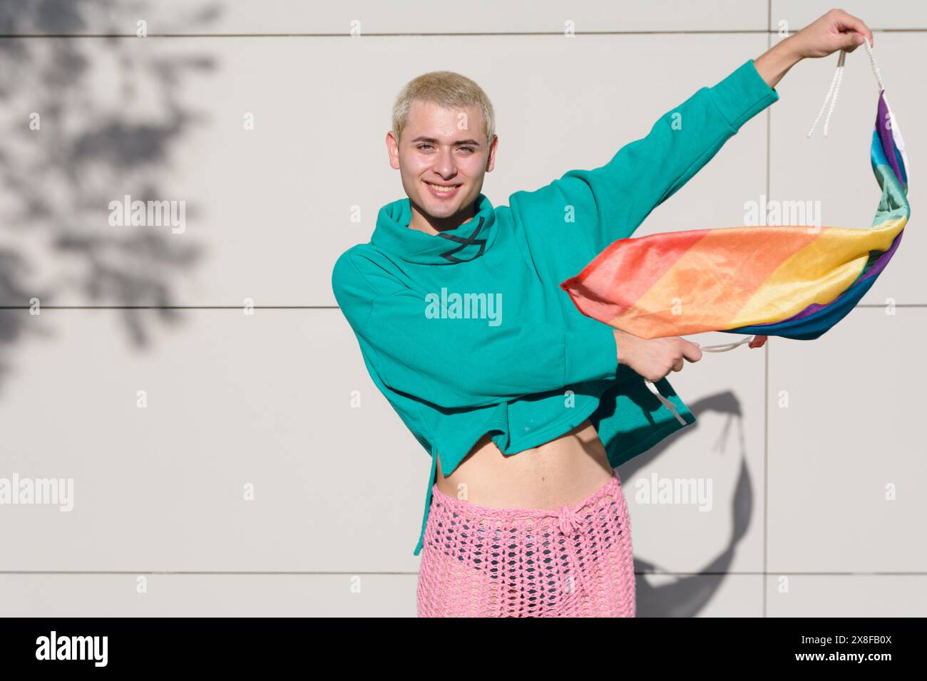 Un jeune homme heureux dans une chemise verte et un pantalon rose en plein air dansant avec son drapeau arc-en-ciel, il est blond et a les cheveux courts, regarde la caméra smili Banque D'Images