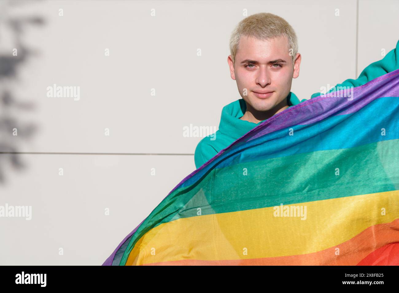 Un jeune homme blond dehors avec son drapeau de fierté debout et heureux. il a les cheveux courts et un sweat à capuche vert regarde la caméra avec détermination et em Banque D'Images