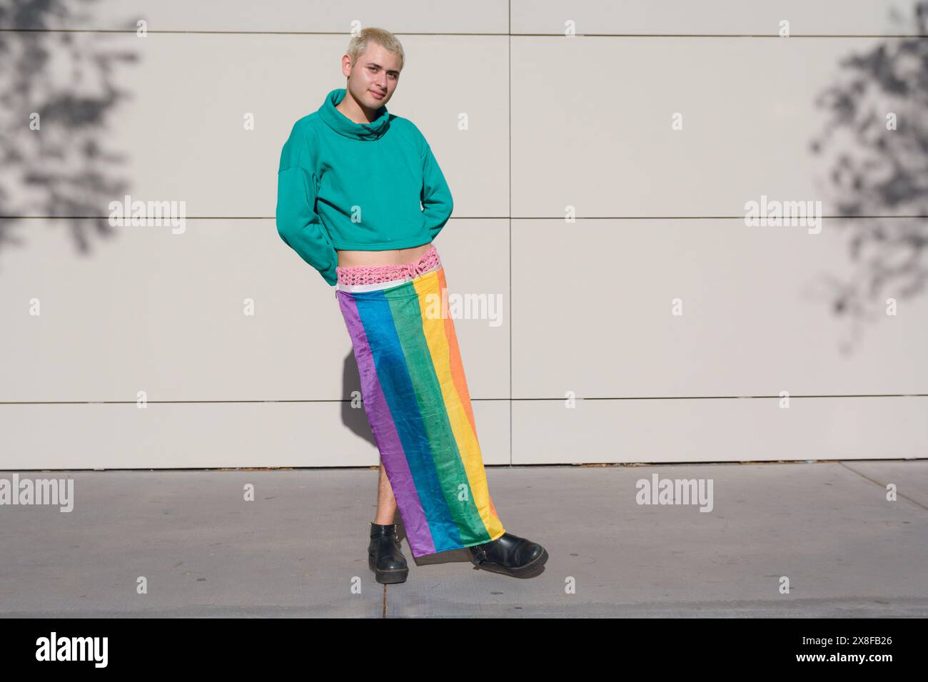 Un homme dans un sweat à capuche vert porte un drapeau de fierté comme une jupe. Il se tient devant un mur à midi, regarde la caméra souriant et posant, copie Banque D'Images