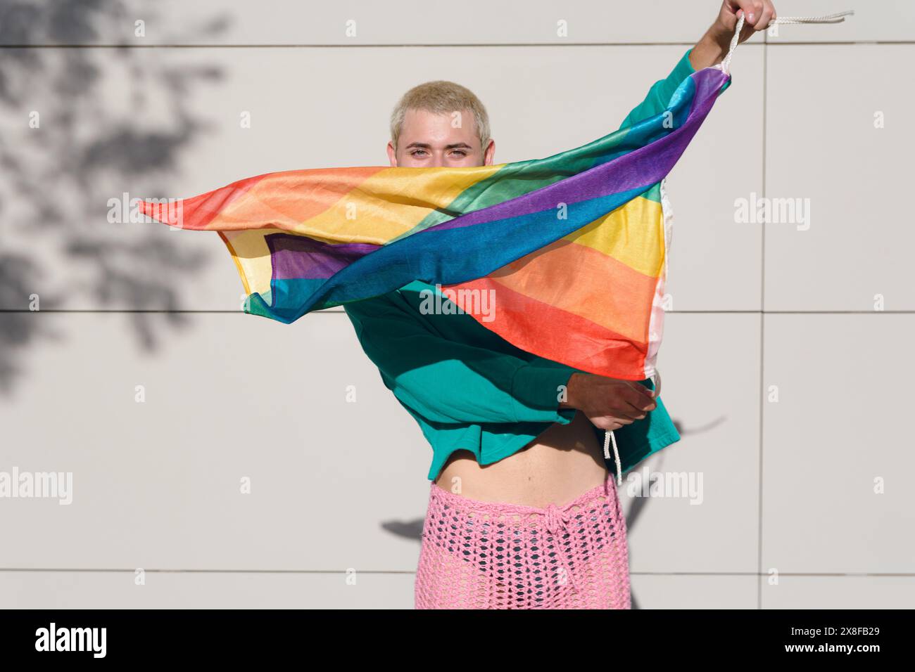 Un jeune homme portant un pull vert et un short en tissu translucide rose tient un drapeau arc-en-ciel. Concept de fierté et de célébration, pose avec le Banque D'Images