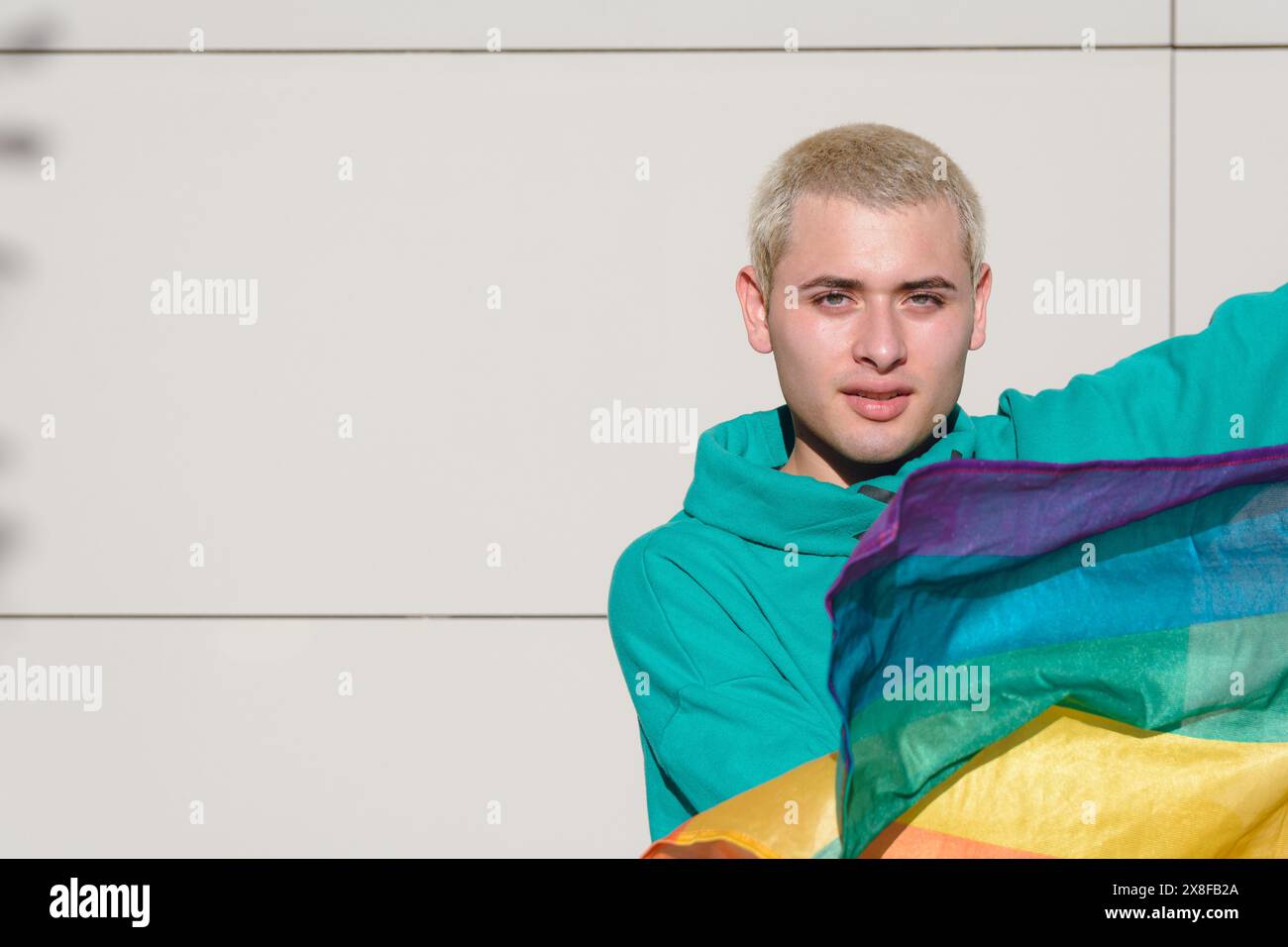 Un jeune homme fier de lui-même agitant un drapeau arc-en-ciel dans sa main. Il porte un sweat à capuche vert. est debout dehors dans une journée ensoleillée en souriant et en regardant Banque D'Images