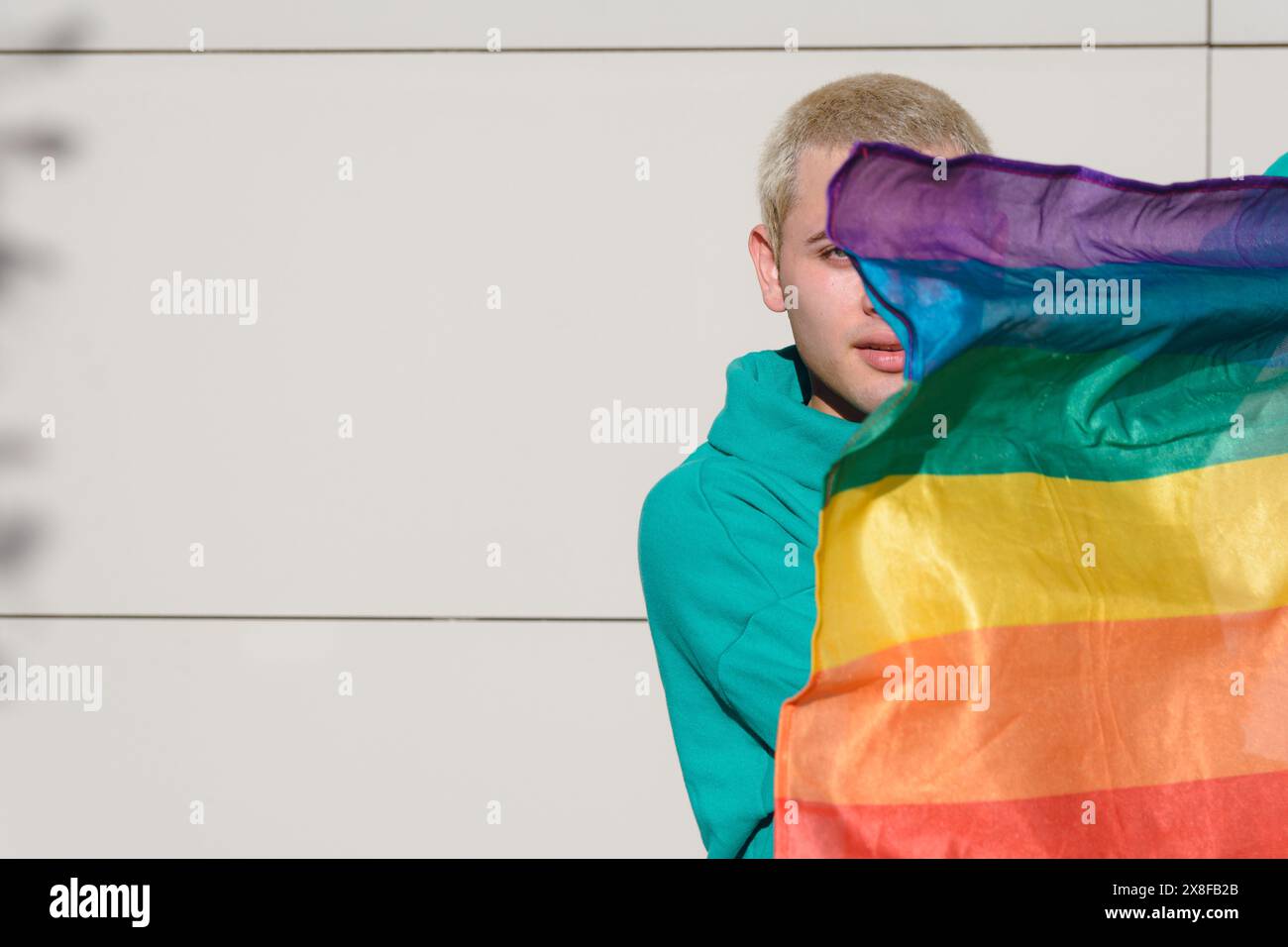 Un jeune homme latin tient un drapeau arc-en-ciel et est partiellement couvert par celui-ci. C'est un homme blond argentin, debout dehors avec un mur sur le dos Banque D'Images