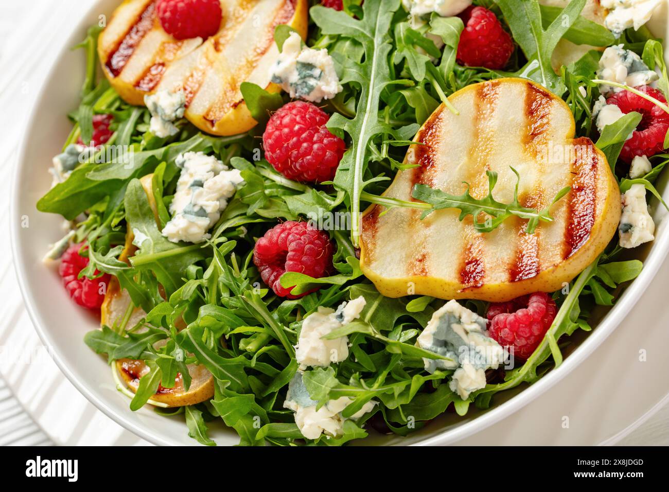 gros plan de salade de poires grillées avec fromage bleu, roquette et framboises fraîches mûres sur une assiette blanche sur une table en bois blanc Banque D'Images