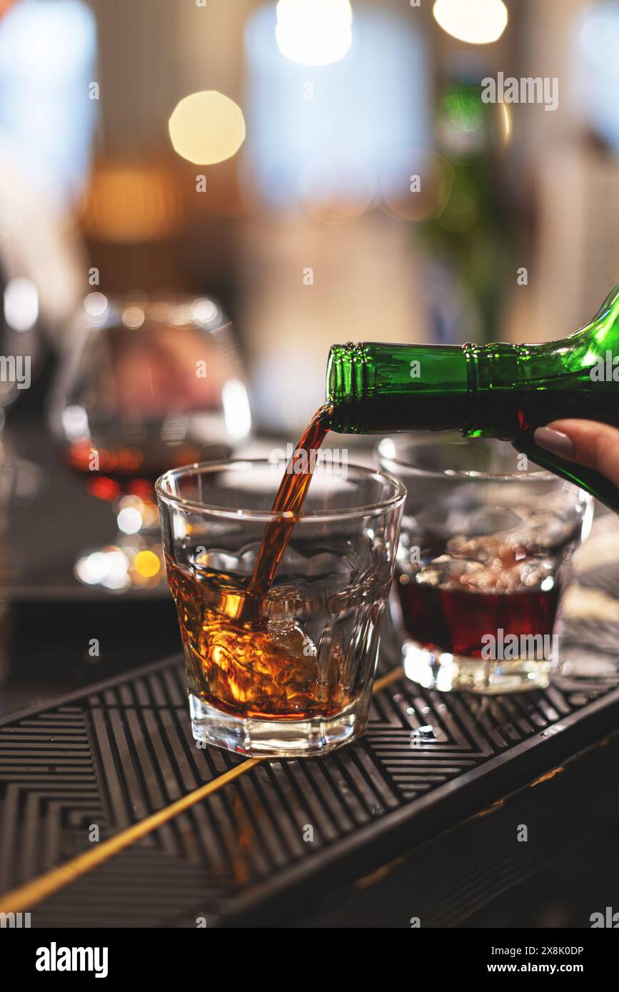 Gros plan d'une main versant du whisky ambré d'une bouteille dans un verre de roches dans un bar avec des lumières bokeh Banque D'Images