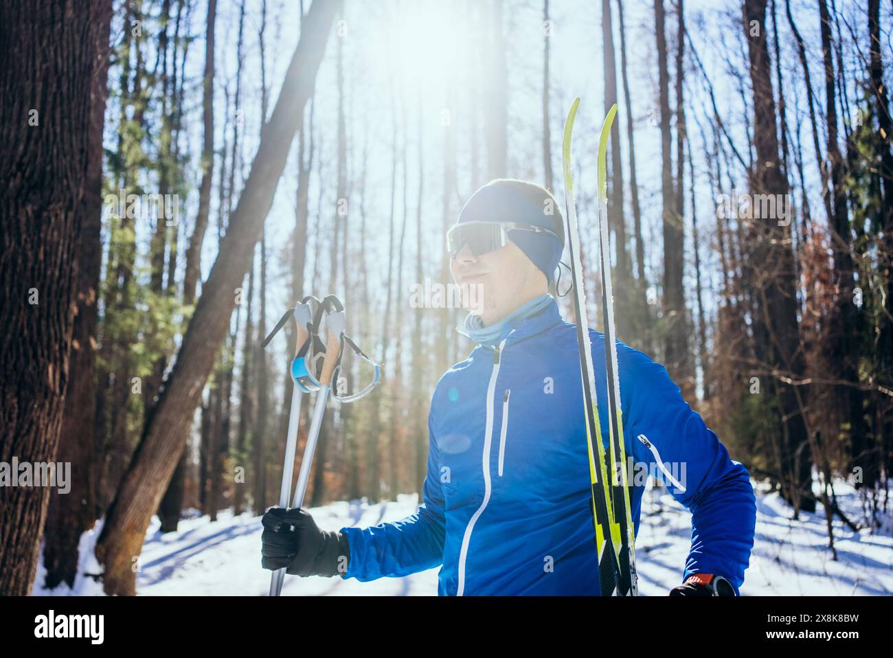 L'homme se tient confiant dans la forêt ensoleillée, prêt pour le ski de fond. Il porte une veste bleue, des lunettes de soleil et un bandeau, tenant des bâtons de ski et des skis. Banque D'Images