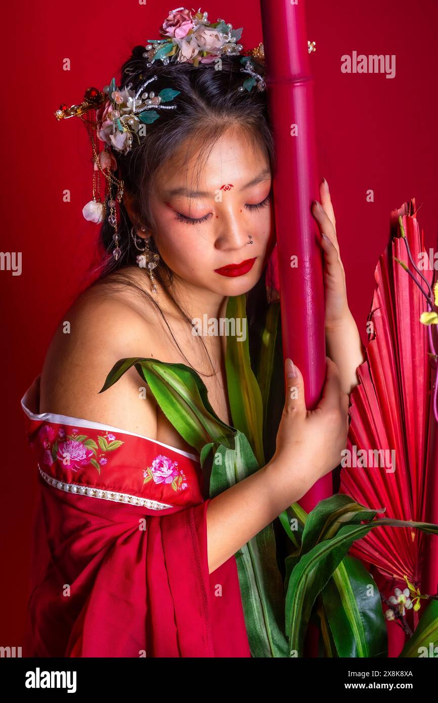 Une femme en robe rouge tient une plante verte. La femme porte un bandeau de fleur et a du rouge à lèvres rouge. L'image a un caractère romantique et féminin Banque D'Images