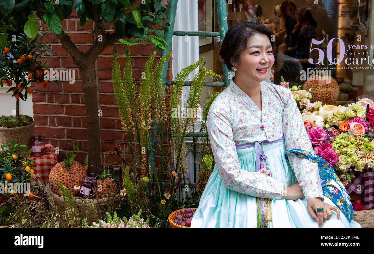 Femme en robe traditionnelle coréenne hanbok assise par un affichage de fleurs souriant avec un fond de mur de briques à Chelsea en fleur 2024 Banque D'Images
