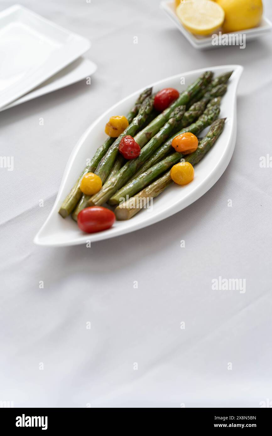 Une source de grandes fibres alimentaires, des asperges vertes cuites au four servies dans une assiette blanche, préparées comme plat d'accompagnement avec des tomates colorées et du citron Banque D'Images