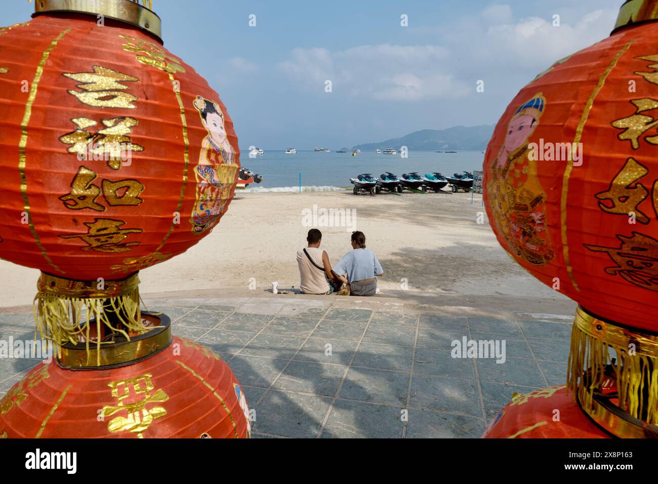 Vu entre deux lanternes chinoises rouges, un couple de touristes est assis sur Patong Beach, Patong, dans le sud de la Thaïlande Banque D'Images