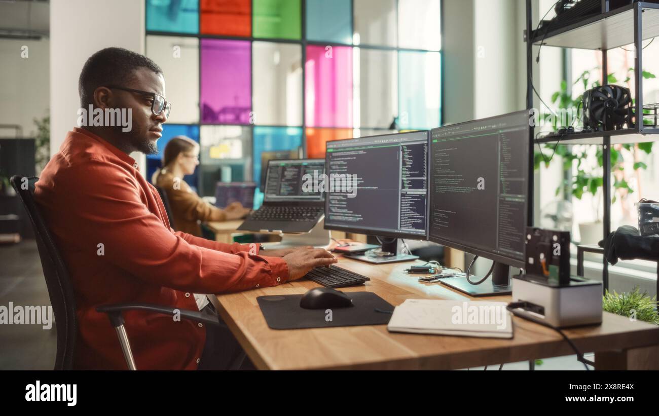 Homme noir écrivant des lignes de code sur un PC de bureau avec deux moniteurs et un ordinateur portable de côté dans un bureau élégant. Développeur professionnel masculin Programming logiciel d'intelligence artificielle pour start-up Company. Banque D'Images