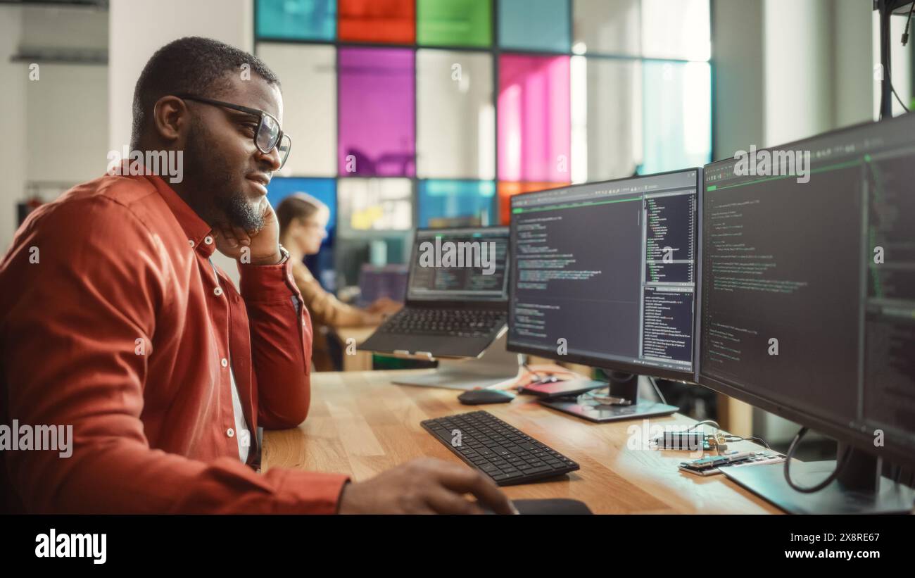 Homme noir écrivant des lignes de code sur un PC de bureau avec deux moniteurs et un ordinateur portable dans un bureau élégant. Développeur professionnel masculin Programming logiciel d'intelligence artificielle pour start-up Company. Banque D'Images