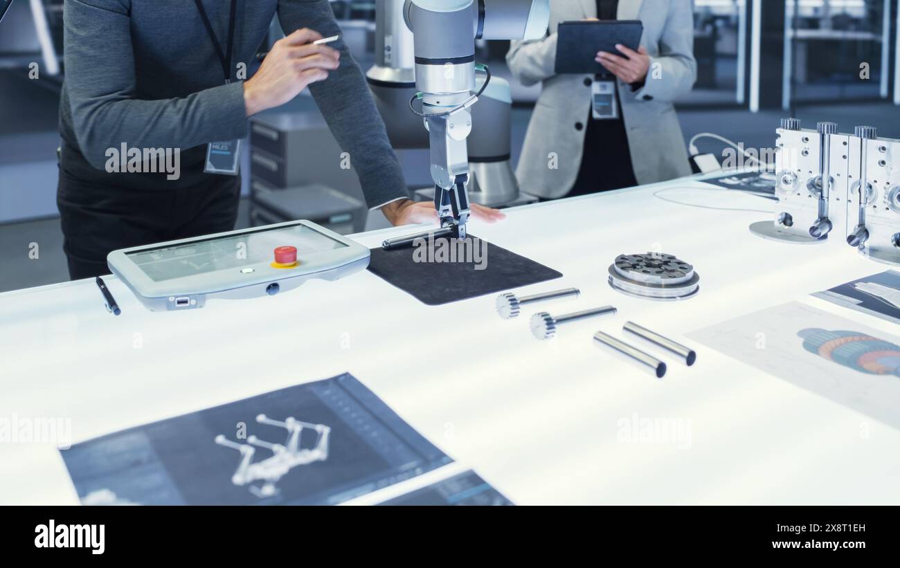 Bras robotique moderne tenant et déplaçant un composant mécanique métallique. Équipe diversifiée d'ingénieurs utilisant un ordinateur tablette pour manipuler et programmer le robot. Banque D'Images