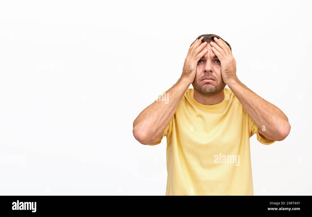 Homme dans une chemise jaune avec ses mains sur la tête, levant les yeux avec une expression très perplexe et déconcertée. isolé sur fond blanc. Banque D'Images