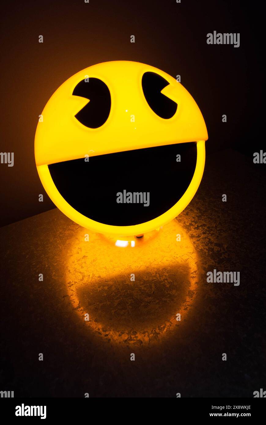 Lampe à visage smiley jaune illuminée projetant une lueur joyeuse et une ombre sur une surface texturée dans une pièce sombre Banque D'Images