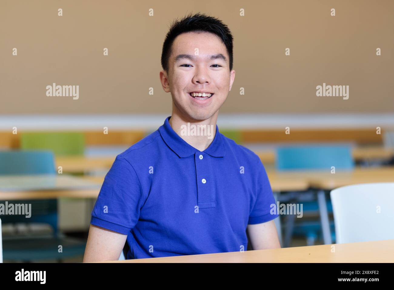 Un jeune étudiant universitaire asiatique en chemise bleue est heureux assis à une table, souriant. Il est à l'intérieur d'une salle de classe d'école. Banque D'Images