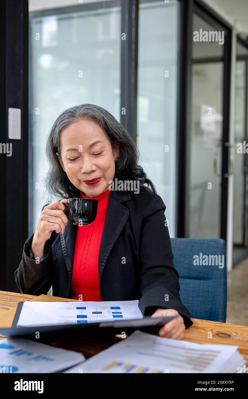 Une femme d'affaires asiatique mature confiante et satisfaite ou un patron féminin sirote un café tout en lisant le rapport d'affaires à son bureau. Banque D'Images