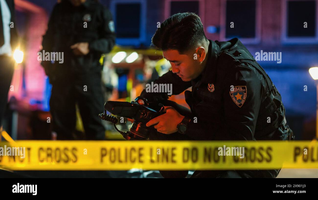 Policier vérifiant la caméra après avoir pris des photos médico-légales des preuves trouvées sur la scène de crime. Jeune policier derrière une bande jaune Don't Cross, résolvant une affaire de meurtre et documentant des indices Banque D'Images