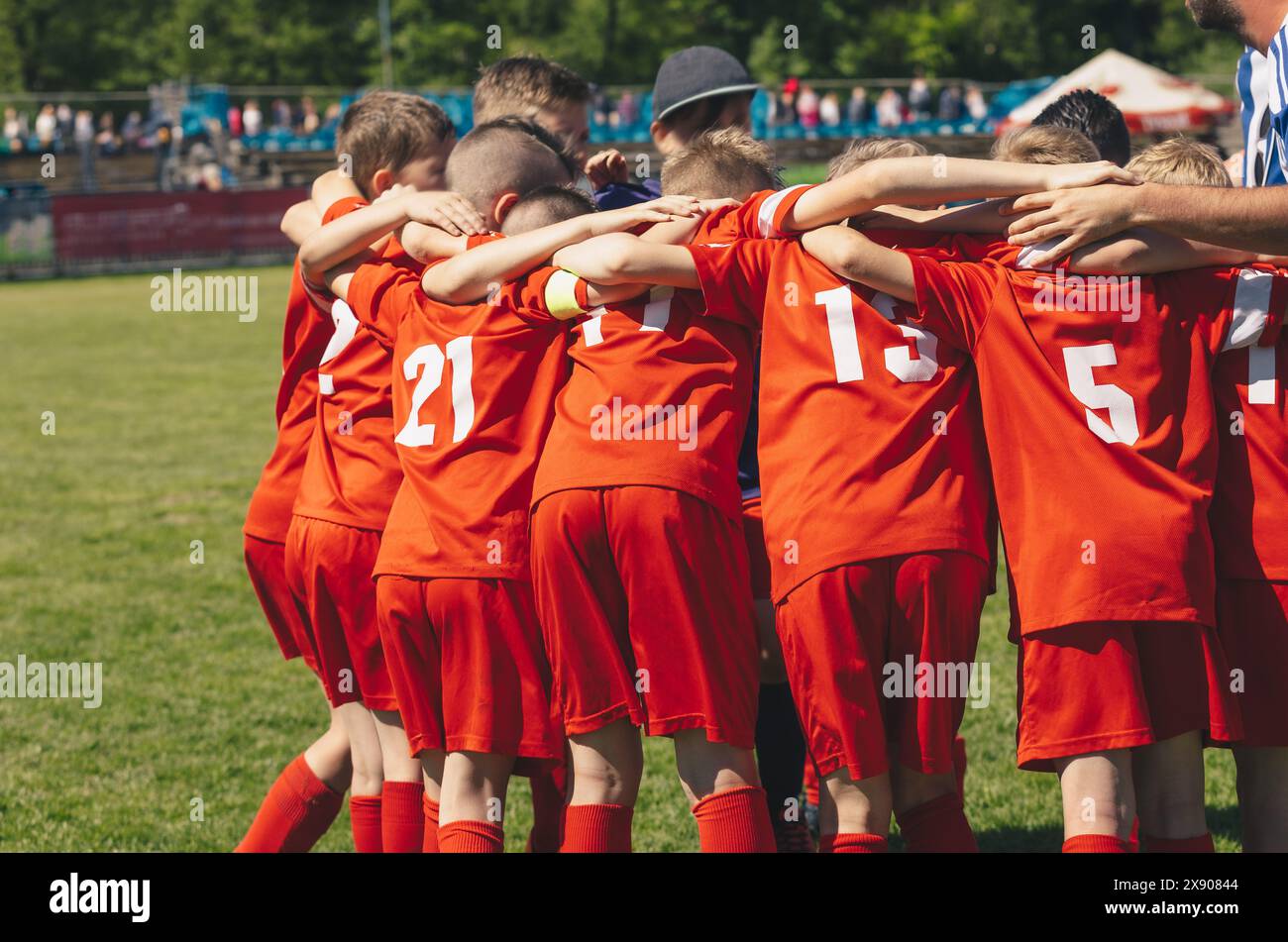 Équipe sportive pour enfants avec entraîneur. Les enfants jouent un match de sport sur le terrain du stade. Écoliers en maillots rouges se blottissant dans une équipe. Parents assis à th Banque D'Images