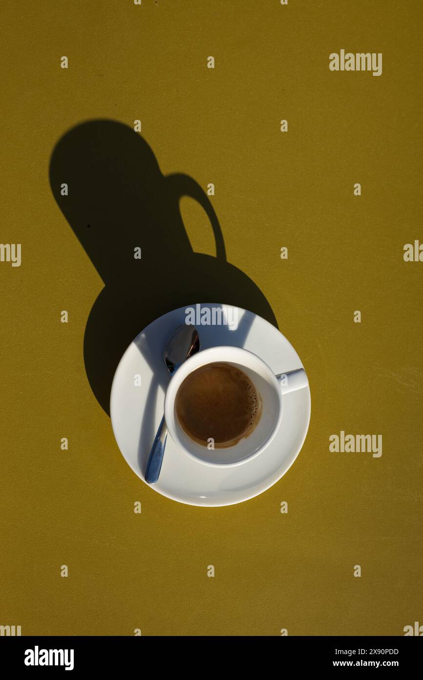 Un café expresso dans une tasse au petit déjeuner dans un café en Italie par une journée ensoleillée avec une table jaune et le soleil projetant une ombre de la tasse et de la soucoupe. Banque D'Images