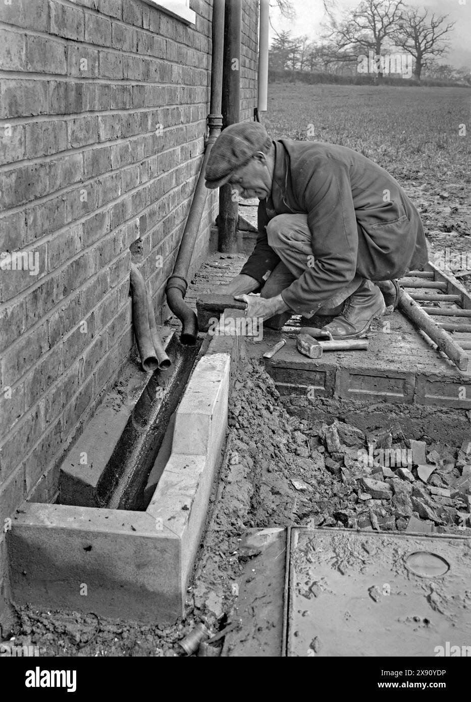 Ouvrier du bâtiment construisant une goulotte ou un canal de drainage extérieur. Le travail a eu lieu dans une maison à Kennel Lane, Fetcham, Leatherhead, Surrey, Angleterre, UK c. 1930. La tuyauterie à cette époque comprenait du plomb (les deux tuyaux sortant à travers le mur) et de la fonte (le plus grand tuyau de descente de la gouttière du toit). Le ravin lui-même est en partie en béton, avec le travailleur finissant la section arrière en brique - une photographie vintage des années 1920/30. Banque D'Images