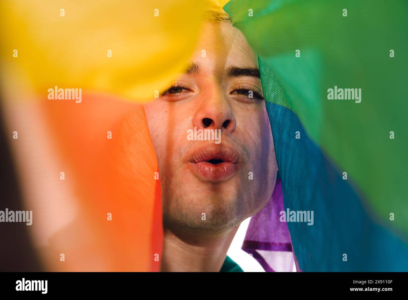 Gros plan de face de Latin Gay Man regardant la caméra souffler un baiser couvert par un drapeau de fierté. Concept d'humour et de légèreté, comme l'homme est ma Banque D'Images