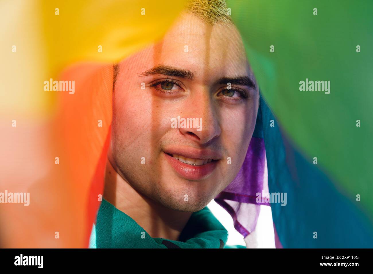 Jeune homme latin gay regardant la caméra souriant et couvert par un drapeau de fierté. Concept de bonheur et fierté dans l'identité homosexuelle de l'homme Banque D'Images