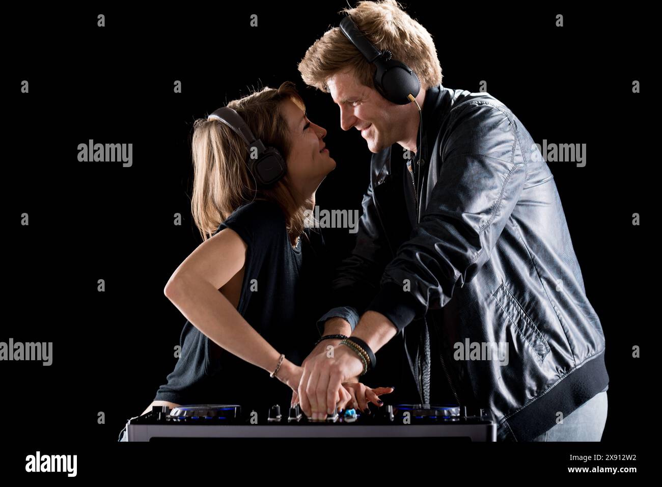 Deux DJ partagent un moment ludique et affectueux tout en travaillant ensemble sur un mixeur. Les deux portent des écouteurs ; la femme se penche avec un sourire, tandis que l'homme, Banque D'Images
