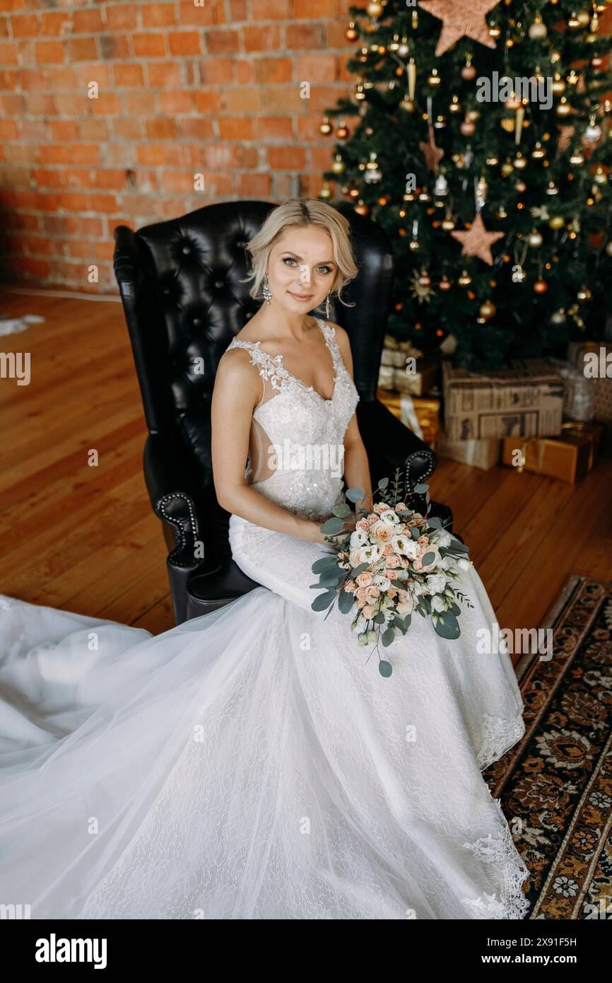 Mariée dans une robe de mariée tenant un bouquet, assise sur une chaise en cuir noir avec un arbre de Noël et des décorations derrière elle Banque D'Images