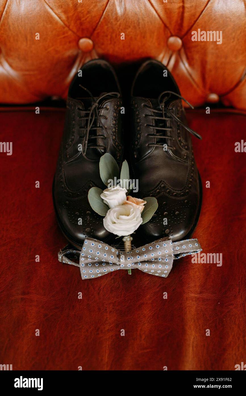 Chaussures de mariage en cuir noir avec une boutonnière florale blanche et un noeud papillon bleu placé sur un canapé en cuir marron Banque D'Images