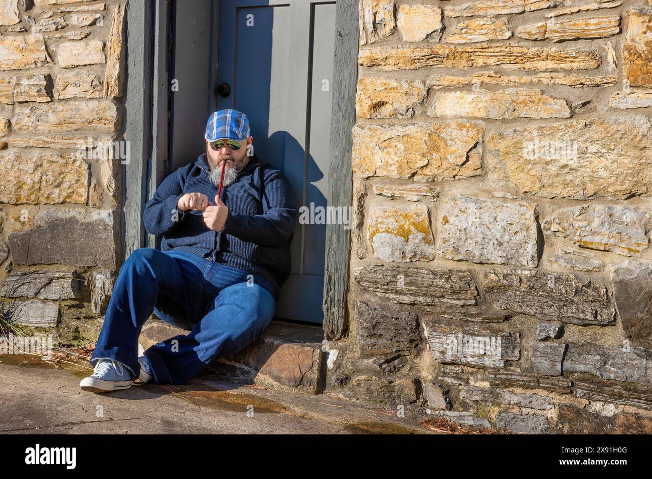 Un homme allume un tuyau tout en étant assis sur une marche devant une porte menant à un bâtiment rocheux. Banque D'Images