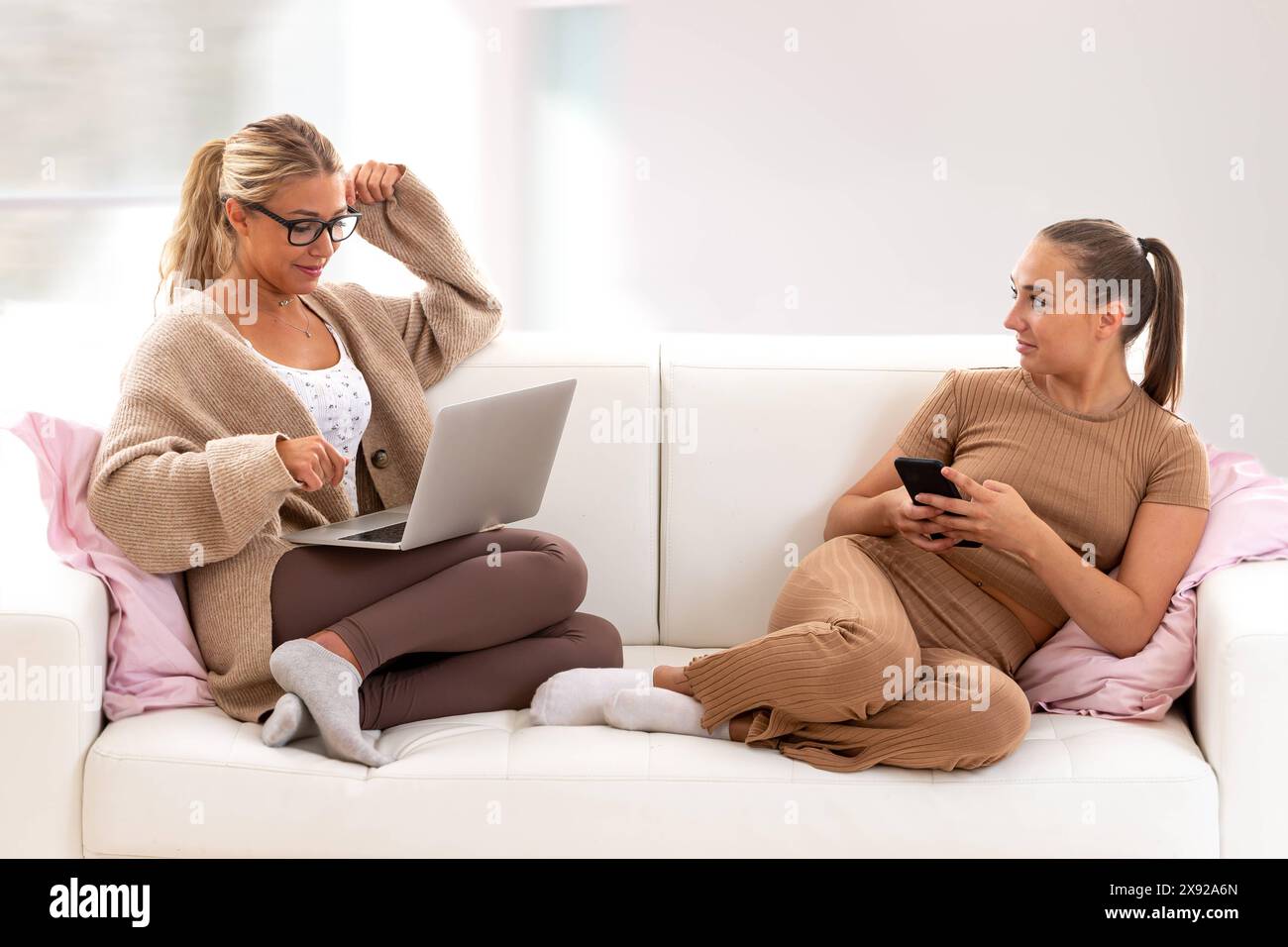 Dialogue décontracté avec ordinateur et smartphone dans les mains. Deux jeunes femmes détendues sur un canapé regardant un site sur Internet. 016644 058 Banque D'Images