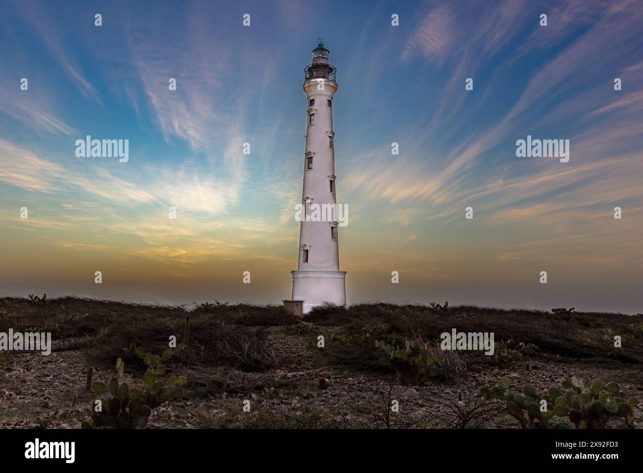 Phare californien d'Aruba au coucher du soleil. Premier plan sombre ; nuages tordus colorés dans le ciel bleu en arrière-plan. Banque D'Images