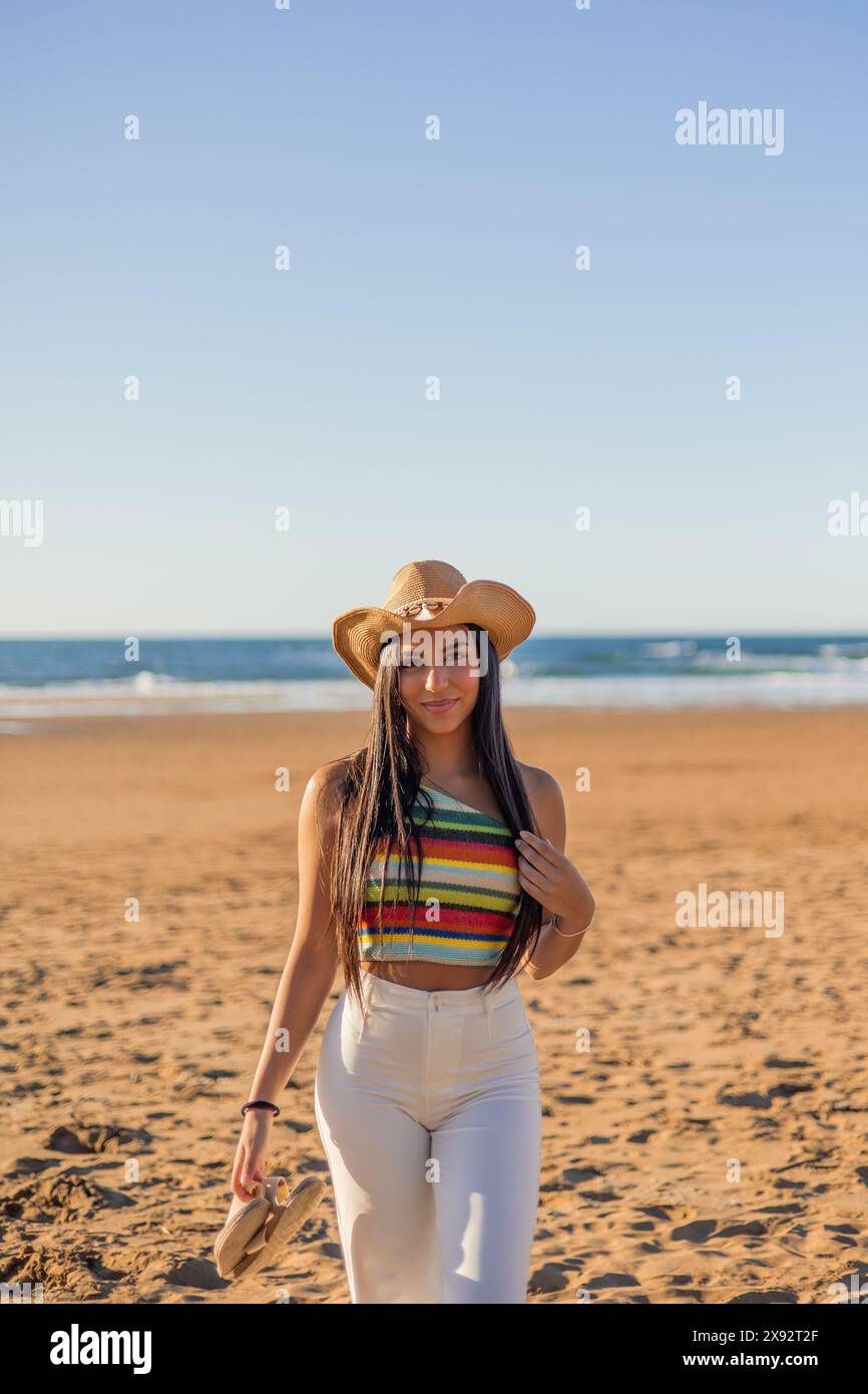 Beauté multiculturelle verticale comme une femme latina prend la pose sur la plage ensoleillée, son chapeau de soleil élégant ajoutant une touche d'élégance à la scène vibrante Banque D'Images