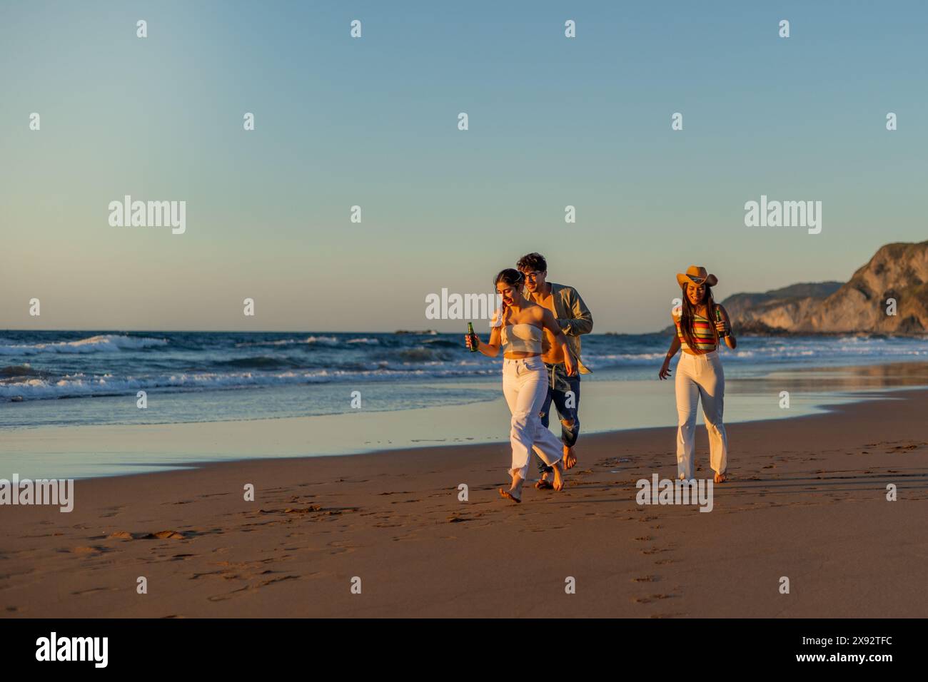 Trois amis hispaniques s'amusant à courir le long du littoral, en dégustant des bières froides pendant un copyspace d'été au coucher du soleil Banque D'Images